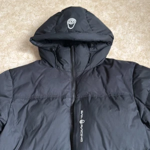 Svart Sail Racing dunjacka med huva - Svart dunjacka från Sail Racing med huva och snygga logodetaljer på bröstet och ärmen. Jackan har dragkedja framtill, praktiska fickor och en cool patch med 'Sail Antarctica Expedition' på ärmen. Perfekt för kalla dagar och har en schysst, lite längre passform.