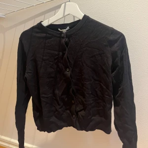 Svart kofta med knappar - En klassisk svart kofta med rund hals och knäppning framtill. Långärmad och i mjukt material, perfekt att slänga över en t-shirt eller linne. Enkel och stilren design som funkar till det mesta.