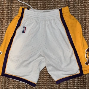 Lakers shorts Mitchell & Ness S - Säljer ett par klassiska Los Angeles Lakers basketshorts från Mitchell & Ness Hardwood Classics. De är vita med gula och lila paneler, Lakers-logga på benet och NBA-märke. Meshmaterial med snörning i midjan, perfekt för basket eller streetwear. Passar M och S