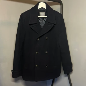 Svart skepparkavaj från Burton Menswear - Klassisk svart skepparkavaj från Burton Menswear London. Jackan har dubbelknäppning med sex knappar, bred krage och två snedställda fickor framtill. Tillverkad i ullblandning med slät fodrad insida. 