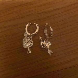 Silverörhängen med hjärta och pil - Säljer ett par snygga örhängen i silverfärg med hängande hjärtan som har blixtformad spricka och pil genom sig. Ringen har små glittriga stenar på framsidan för extra bling. Perfekt accessoar för att sticka ut lite extra. Aldrig använda