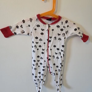 Vit pyjamas med svart mönster och röda detaljer - Söt vit pyjamas med svarta mönster av träd och djur, röda muddar och krage. Dragkedja framtill för enkel påklädning. Mjukt bomullsmaterial och hel täckning för både armar och ben, perfekt för chill dagar hemma.