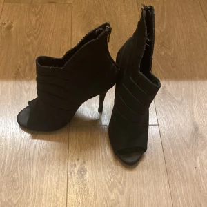 Svarta stövletter med öppen tå från BIANCO - Svarta stövletter från BIANCO med hög klack och öppen tå. Snyggt ribbade detaljer på ovansidan och dragkedja baktill. Tillverkade i mocka för en lyxig känsla. Perfekt för dig som vill ha en edgy och stilren look.