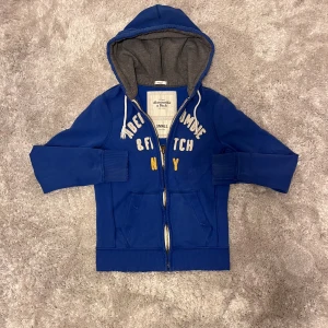 Blå hoodie från Abercrombie & Fitch - Snygg blå hoodie från Abercrombie & Fitch med broderad text i vitt och gult på bröstet. Den har ett litet hål i ärmen. Hör av er vid frågor!
