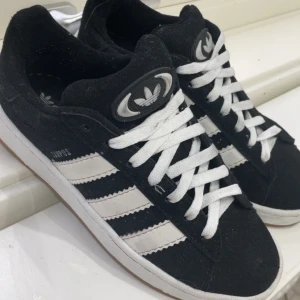 Adidas Campus svarta skor - Svarta Adidas Campus sneakers med klassiska vita ränder på sidorna och vit logga på plösen. Skorna har snörning och en platt gummisula i brunt. Ovandelen är i mocka och insidan är mjuk för extra komfort. Perfekt för dig som gillar retrostil och streetwear. Har bara haft på mig de en gång och köpte de i juli 2025, priset går o diskuteras 