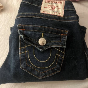 True Religion Joey Low Rise Flare Jeans - Mörkblå jeans från True Religion, modell Joey Low Rise Flare. Snygga kontrastsömmar i gult och orange, ikoniska ficklock med silverfärgade knappar och klassisk logga bak. Bootcut/flare passform och låg midja. Perfekta för dig som gillar statement-jeans! Jag har kortat ner dem hos en skräddare och jag är ca 155 och de passar bra i längd 