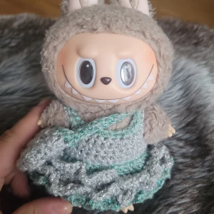 Handvirkad silvergrå labubu klänning  - Supersöt handvirkad klänning i silvergrått och mintgrönt garn med glittriga detaljer. Klänningen har volangkant och matchande liten väska. Perfekt för att ge dockan en unik och gullig look.