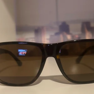 Polariserade Armani Solglasögon  - Snygga solglasögon från Armani med brunmelerad båge och bruna polariserade glas. Rektangulär form och diskret logga på skalmen. Perfekt accessoar för att lyfta din stil i sommar. Äkta Köpta på synoptik 