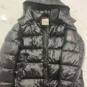 Svart Moncler pufferjacka med huva - Svart pufferjacka från Moncler med glansig finish, huva och klassisk logga på ärmen. Jackan har dragkedja framtill och snedställda fickor med dragkedja. Perfekt för dig som vill ha en snygg och varm jacka med street vibe. Dragkedjan kan kännas svårt att få upp. Dock så beror det på att jackan inte har används och där av priset ! 