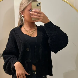 Svart stickad kofta med stora knappar - Säljer en svart stickad kofta med oversized passform och stora, runda knappar framtill. Koftan har breda, ribbade muddar och långärmade ballongärmar som ger en cool och avslappnad vibe. Perfekt att slänga över en topp för en snygg lager-på-lager-look.