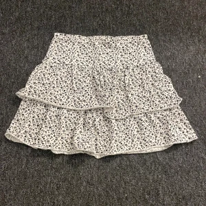 Vit blommig volangkjol från Lindex - Supersöt kort kjol från Lindex med volanger och blommigt mönster i svart på vit botten. Kjolen har en bred midja och är gjord i mjuk bomull, perfekt för dig som gillar en feminin och chill stil. Passar dig som är ca 14 år eller äldre.