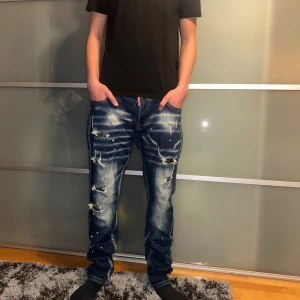 Blå d squared jeans med färgstänk - Säljer ett par blå d squrered2 med coola slitningar och färgstänk i rött och vitt. 44cm i bredden. Jeansen har en straight passform, klassiska fem fickor och snygga detaljer som ger en unik streetkänsla. Perfekt för dig som vill sticka ut med din stil.