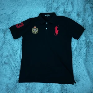 Svart piké från Polo Ralph Lauren - ” Passar även folk med S”   röd broderad logga på bröstet, brittiskt emblem i guld och rött samt stor röd siffra 3 på ärmen. 