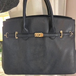 Mörkblå handväska med gulddetaljer - Stilren mörkblå handväska i skinnimitation med guldfärgade metalldetaljer och klassiskt lås framtill. Väskan har två rejäla handtag och en praktisk dragkedjeficka på baksidan. Perfekt för dig som vill ha en tidlös och elegant accessoar.