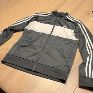 Adidas svart och grå track jacket - Snygg Adidas track jacket i grått med bred vit panel över bröstet och ryggen. Klassiska vita ränder längs ärmarna och Adidas-logga på bröstet. Jackan har hel dragkedja, ribbade muddar och är tillverkad i mjuk polyester.Fläckar förekommer på det vita men syns bara om man tittar noga 