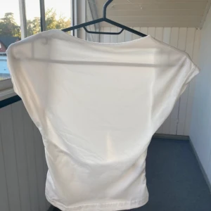 Vit topp med öppen rygg - Säljer en vit topp i mjukt material med bred halsringning och öppen rygg. Toppen har korta ärmar och en lätt insvängd midja. Perfekt för dig som gillar en enkel men ändå snygg stil. 