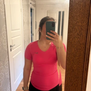 Rosa tränings T-shirt  från Nike - Säljer en snygg rosa träningstopp från Nike med v-ringning och korta ärmar. Toppen har en tight passform och är gjord i ett mjukt syntetmaterial som andas bra vid träning. Diskret Nike-logga på bröstet. Perfekt för gymmet eller löprundan! Aldrig använd, så mycket bra skick. Djur och rökfritt hem. Vid snabb affär kan jag tänka mig att sänka priset. 