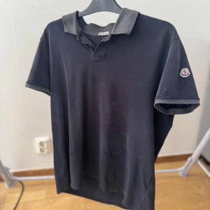 Svart pikétröja från Moncler - Säljer en svart pikétröja från Moncler med grå krage och ärmslut. Klassisk Moncler-logga på vänster ärm. Tröjan har korta ärmar och är tillverkad i bomull för en skön och luftig känsla. Perfekt för dig som gillar stilrena och tidlösa plagg.