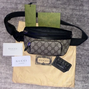 Gucci beige magväska med GG-mönster - Säljer en snygg magväska från Gucci i beige och svart med klassiskt GG-mönster. Väskan har svarta detaljer i skinn, justerbart band och dragkedjor både fram och bak. Perfekt för att bära det viktigaste och ge outfiten en lyxig touch.
