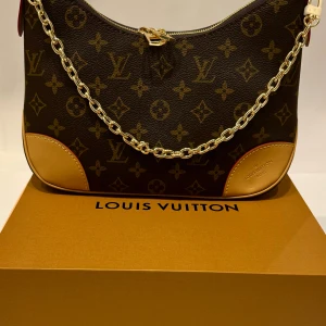 Louis Vuitton monogram axelväska - Snygg axelväska från Louis Vuitton med klassiskt monogrammönster i brunt och beige. Väskan har guldfärgad kedja och detaljer i ljust skinn. Perfekt för dig som vill ha en ikonisk och trendig accessoar med lyxig känsla.