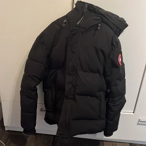 Svart pufferjacka från Canada Goose - Svart pufferjacka från Canada Goose med ikonisk patch på ärmen, stor huva och dragkedja framtill. Jackan har en clean och chunky look med breda, quiltade sektioner och praktiska fickor. Perfekt för kalla dagar när du vill hålla stilen.