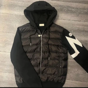 Svart Moncler pufferjacka med huva - Svart pufferjacka från Moncler med huva och dragkedja framtill. Jackan har quiltad front i blankt syntetmaterial, stickade ärmar med vit zigzag-mönster och Moncler-logga på ena armen. Snygga detaljer som snörning vid huvan och silverfärgad dragkedja. Priset är inte hugget i sten kontakta vid intresse! Storlek L passar M också.