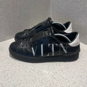 Valentino skor - Snygga svarta sneakers från Valentino Garavani med stor VLTN-logga i vitt på sidan och vit häl. Skorna har snörning, platt sula och är tillverkade i skinn. Perfekt för dig som gillar exklusiv streetstyle och vill sticka ut med designer på fötterna.