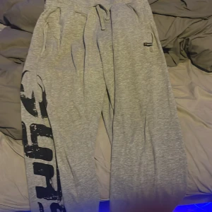 Grå mjukisbyxor från Gasp, storlek L - Säljer ett par grå mjukisbyxor från Gasp i storlek L. Byxorna har snörning i midjan, fickor och ett stort svart tryck längs ena benet. Sköna och loose passform, perfekta för chill eller träning. Materialet är mjukt och känns som bomull.