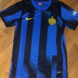 Inter Milan blå-svart matchtröja Nike - Inter Milan 2023/24 hemma fotbollströja från Nike med blå och svarta digitalt randiga paneler, gul Nike-logga och klubbmärke på bröstet. Kortärmad modell i lätt och ventilerande material, perfekt för match eller träning.