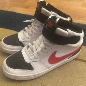 Nike Air Force 1 High vit/svart/röd - Säljer ett par Nike Air Force 1 High sneakers i vitt skinn med svarta och röda detaljer. Skorna har klassisk röd swoosh, svart tå och häl, samt svart kardborreband vid ankeln. Tjock vit sula och snörning framtill. Perfekta för dig som gillar retrostil och streetwear.