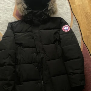 Canada Goose Wyndham  - Säljer en svart dunjacka från Canada Goose med pälskantad huva och klassisk patch på ärmen. Jackan är quiltad, har dragkedja och knappar framtill samt flera praktiska fickor. Perfekt för kalla vinterdagar och riktigt snygg streetstil.