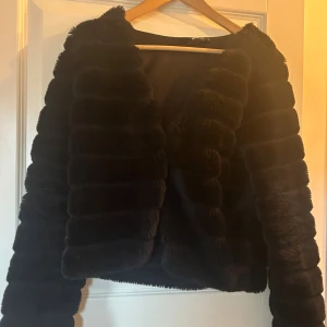 Svart pälsjacka med ribbad look - Säljer en svart pälsjacka med horisontella ribbade partier. Jackan är kortare i modellen och har långa ärmar. Perfekt för dig som vill ha en snygg och trendig look med lite extra attityd. Supermjuk och fluffig känsla.
