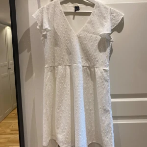 Vit spetsklänning från Vero Moda - Superfin vit kortklänning från Vero Moda med v-ringning och volangärmar. Klänningen har broderad spets över hela tyget och en lätt utställd kjol. Perfekt för sommarens alla tillfällen och ger en fräsch och romantisk vibe.