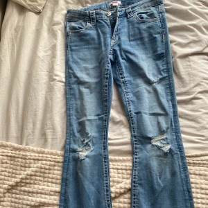 Blå bootcut jeans från Gina Tricot - Snygga ljusblå jeans från Gina Tricot med bootcut passform och slitna detaljer på benen. Baksidan har broderade fickor med coolt mönster. Jeansen har klassisk femficksdesign och knapp med blommönster. Perfekt för dig som gillar en avslappnad och trendig stil.