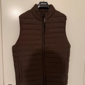 Brun dunväst från Jack & Jones  - Mörkbrun quiltad dunväst från Jack &jones med hög krage och dragkedja framtill. Västen har en stilren, rak passform och horisontella sömmar som ger en klassisk look. Perfekt att slänga över en hoodie eller tröja när det är kyligt ute.