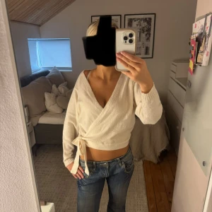 Vit croppad stickad tröja med knyt - Säljer en vit croppad stickad tröja med knyt i sidan och v-ringad hals. Toppen har långa ärmar och är tillverkad i ett mjukt stickat material som känns skönt mot huden. Storlek S. 