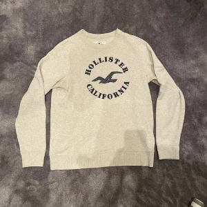 Hollister sweatshirt - Snygg grå sweatshirt från Hollister med stort svart tryck på bröstet där det står 'Hollister California' och en ikonisk fågel. Tröjan har rund hals, ribbade muddar och är tillverkad i mjuk bomullsmix. Otroligt populär, storlek M och fint skick!! Vid fler frågor är det bara att höra av sig!