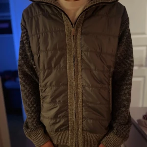 Grå quiltad jacka med stickade ärmar - Snygg grå jacka med quiltad framsida och stickade ärmar. Jackan har dragkedja och ribbade muddar. Perfekt för kyligare dagar. Samt väldigt skön eftersom det är mjukt tyg i!