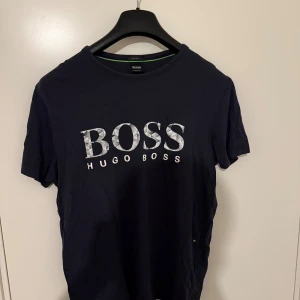 Mörkblå Hugo Boss t-shirt med logga - Snygg mörkblå t-shirt från Hugo Boss med stor vit logga framtill. Klassisk rund halsringning och korta ärmar. Tillverkad i mjuk bomull som känns skön mot huden. Perfekt för dig som gillar stilrena och trendiga plagg med tydlig branding. Äkta såklart 