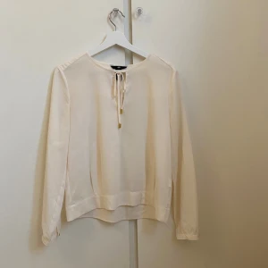 Vit blus med gulddetaljer - Säljer en stilren vit blus från H&M med långärmade ärmar och rund halsringning. Framtill finns ett snyggt knyt med guldfärgade detaljer. Blusen har en rak passform och är tillverkad i ett lätt, mjukt material som ger en skön känsla.