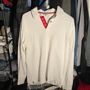 Polo burberry  - Vit långärmad pikétröja från Burberry i storlek XXL men som passar M. Klassisk krage och knäppning med snygga röda detaljer och Burberry-logga på insidan. Tillverkad i bomull, perfekt för dig som gillar stilrena och exklusiva plagg.