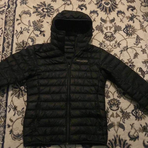 Svart dunjacka Peak Performance - Svart dunjacka från Peak Performance med huva och dragkedja framtill. Jackan är quiltad med horisontella sömmar och har en stilren look. Perfekt för kalla dagar och enkel att matcha med det mesta.