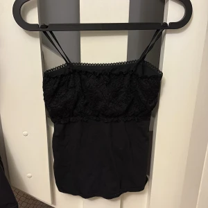 Svart spetsdetaljerad linnetopp - Svart linnetopp med tunna axelband och spetsdetaljer upptill. Toppen har en mjuk, flowy passform och ärmlös design. Spetsen ger en extra touch av stil och kanten har en söt dekorativ kant. Tröjan är i storlek xs/s.