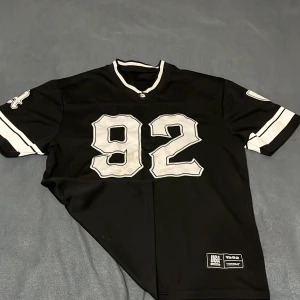 Svart Los Angeles fotbollströja 92 - Snygg svart fotbollströja från USA Sports med stora vita siffror 92 på både fram- och baksidan samt texten LOS ANGELES på ryggen. Tröjan har vita detaljer på ärmar och krage, samt broderade märken på ärmarna. Perfekt för dig som gillar sportig streetstyle.