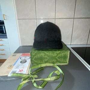 Svart keps från Gucci med GG-mönster - Säljer en svart keps från Gucci i storlek M. Kepsen har det klassiska GG-monogrammönstret ton-i-ton och en grön-röd rand på sidan. Tillverkad i bomullsmix med justerbar rem baktill. Kommer med originalkartong och Gucci-band.