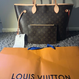 Louis Vuitton Neverfull Monogram Tote - äkta, oanvänd  - Säljer en klassisk Louis Vuitton Neverfull handväska i brunt monogrammönster med beige och röda detaljer. Väskan är rymlig med randigt innerfoder och har en tillhörande clutch med dragkedja. Tillverkad i slitstarkt skinn och canvas, perfekt för dig som vill ha en ikonisk och snygg väska.