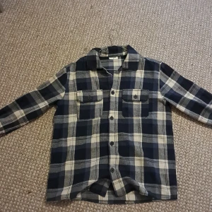 Rutig blå skjorta från Kappahl - Säljer en rutig skjorta från Kappahl i storlek 158. Skjortan har ett klassiskt blått och vitt rutmönster, två bröstfickor med knappar och långa ärmar. Perfekt för dig som gillar avslappnad stil och vill ha något som sticker ut lite extra.