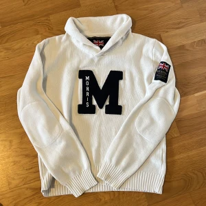 Vit stickad tröja från Morris - Vit stickad hoodie från Morris med stor svart M-applikation på bröstet. Tröjan har ribbade muddar, förstärkta armbågar och en skön krage. Perfekt för kallare väder, fint skick och storlek S, hör av dig vid några frågor!