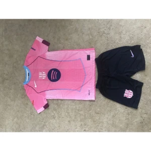 Barça rosa fotbollströja Lamine Yamal - Säljer en FC Barcelona fotbollströja i rosa med blå detaljer och tryck, Lamine Yamal #10 på ryggen. Tröjan har korta ärmar, klubbmärke och sponsorloggor. Matchande svarta shorts med klubbmärke och Nike-logga ingår. Perfekt för dig som älskar Barça!