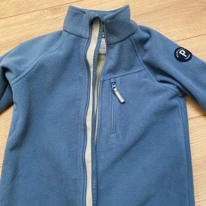 Blå fleecejacka Polarn O. Pyret 122 Didrickson strl 110 - Mysig blå fleecejacka från Polarn O. Pyret med hög krage och dragkedja framtill. Jackan har en bröstficka med dragkedja och ett snyggt PO.P-märke på ärmen. Perfekt för lager-på-lager under kyliga dagar.